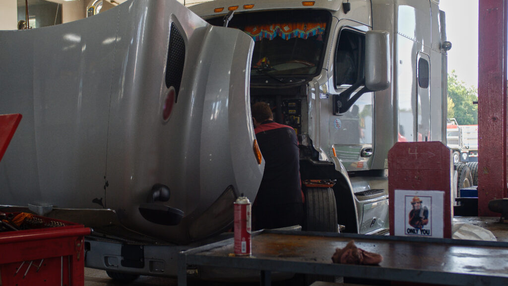 Semi truck with its hood open and a person servicing the engine area within a garage bay.